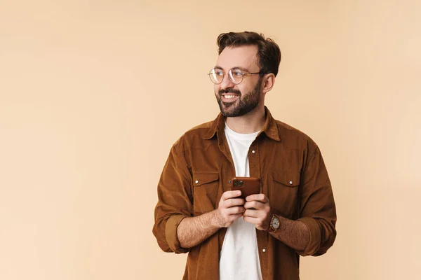ベージュ地の上に立ち 携帯電話を使用して カジュアルな服を身に着けている陽気な若い魅力的な髭の男の肖像画 — ストック写真