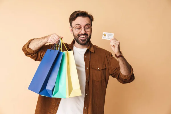幸せな笑顔若い未剃毛男隔離上ベージュ壁背景保持クレジットカードとショッピングバッグ — ストック写真