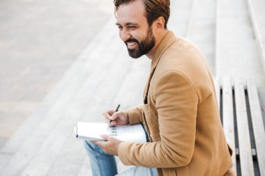 Ceketli yakışıklı yetişkin bir adamın elinde kağıt belgelerle dışarıda bankta otururken uyanması.