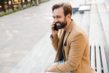 Ceketli, dışarıda bankta otururken akıllı telefondan konuşan yakışıklı bir yetişkin resmi.