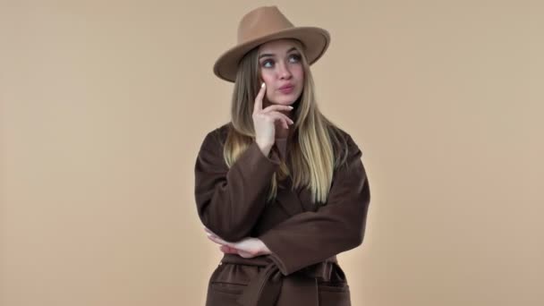 一位头戴帽子 身穿外套的有思想的年轻女子在工作室里想出了一个关于米色背景的主意 — 图库视频影像