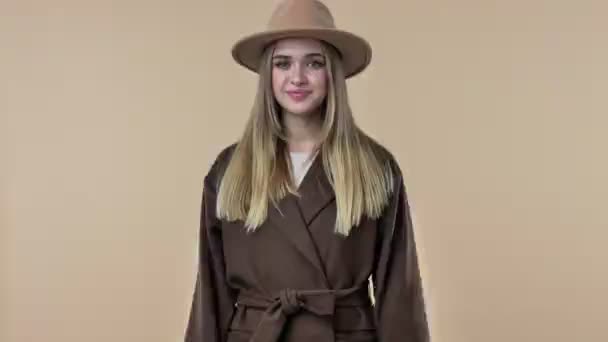 帽子とコートを被った若い笑顔の正の女性がスタジオでベージュの背景の上に隔離されたカメラを指しています — ストック動画