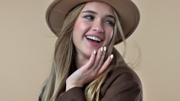 帽子とコートを身に着けている美しい笑顔の若い女性のクローズアップビューは スタジオでベージュの背景に隔離されたポーズをとっています — ストック動画