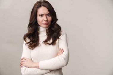 Portrait of an attractive upset young woman with long brunette hair wearing sweater standing isolated over gray background, arms folded clipart