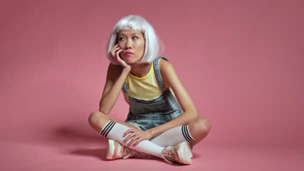 年轻的认真的亚洲女孩穿着白色假发 孤零零地坐在粉红的墙背上 — 图库视频影像