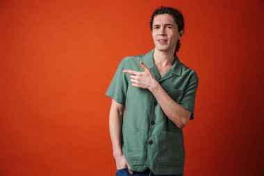 Tedirgin olmuş yakışıklı bir adam, kameraya bakıyor ve turuncu arka planda izole edilmiş bir şekilde işaret ediyor.