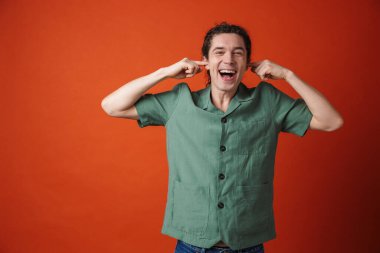 Turuncu arka planda gülen ve kulaklarını tıkayan mutlu yakışıklı bir adam görüntüsü.