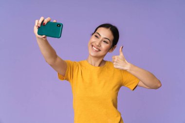 Neşeli, genç, güzel sporcu kadın menekşe arka planda izole bir şekilde duruyor, selfie çekiyor, baş parmağını kaldırıyor.