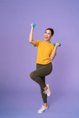 Menekşe arka planında izole bir şekilde duran, halterlerle egzersiz yapan neşeli, genç ve güzel bir kadın.