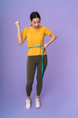 Menekşe arka planında izole bir şekilde duran, belinde bant tutan, başarıyı kutlayan neşeli, genç ve güzel bir kadın.