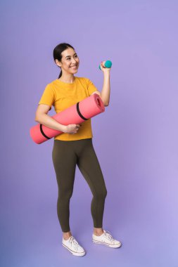 Menekşe arka planında izole bir şekilde duran, halteri kaldırırken spor minderini taşıyan neşeli, genç ve güzel bir kadın.