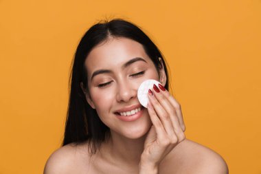 Yüzünü temizleyen ve sarı arka planda izole edilmiş pamuklu ped ile makyaj yapan genç yarı çıplak bir kadının güzel portresi.