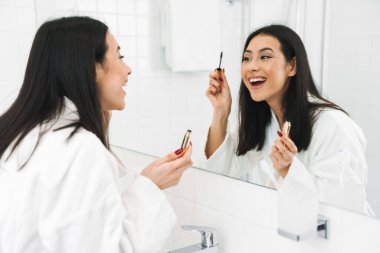 Evde, banyoda gülümseyen neşeli bir genç kadının görüntüsü kirpikleri için makyaj maskarası sür..