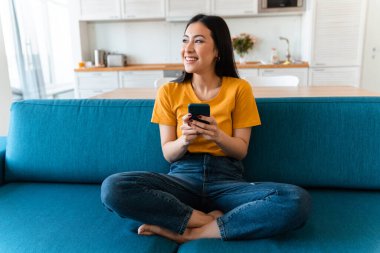 Evdeki kanepede cep telefonuyla gülümseyen güzel bir esmer kadının fotoğrafı..