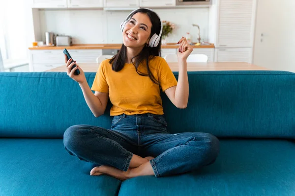 照片中的一个漂亮而积极的 乐观的黑发女子 她坐在家里沙发上 一边听着音乐 一边拿着手机 — 图库照片