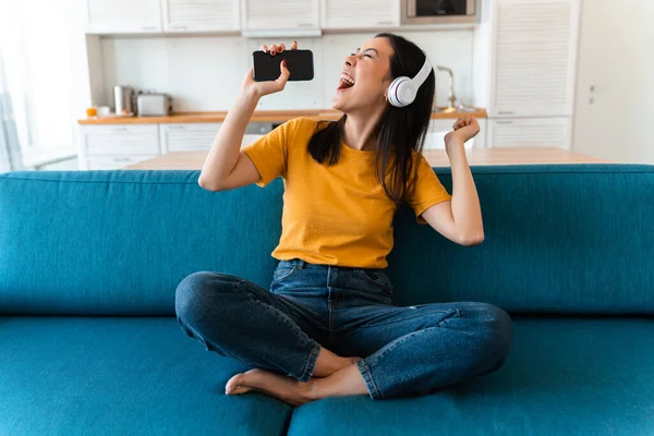 照片上是一位漂亮的 情绪化的 快乐的黑发女子 她坐在家里沙发上 一边听着音乐 一边拿着手机唱歌 — 图库照片
