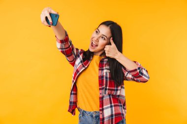 Sarı arka planda izole edilmiş cep telefonunda el hareketi yapan ve selfie çeken komik bir kadın resmi.