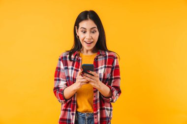 Ekose gömlekli, sarı arka planda izole bir şekilde cep telefonunda yazan şaşırmış çekici bir kadın.