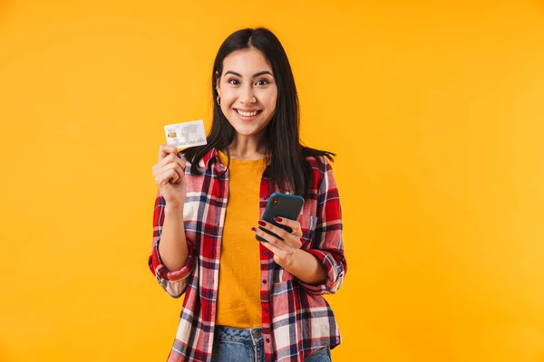 クレジットカードを持っている幸せな魅力的な女性のイメージと黄色の背景に隔離された携帯電話を使用して — ストック写真