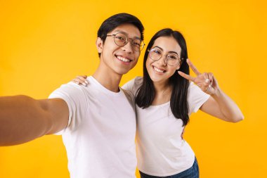Çok uluslu mutlu bir kadın ve erkeğin selfie çekip sarı arka planda izole edilmiş barış işareti yapmaları.
