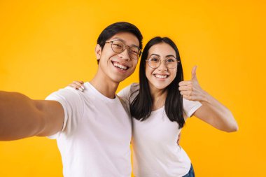 Mutlu çokuluslu bir çiftin selfie çekmesi ve sarı arka planda izole edilmiş barış işareti göstermesi.