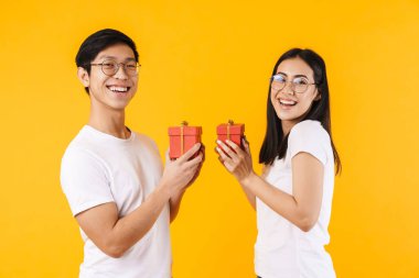 Çok uluslu komik bir çiftin hediye kutuları taşıdığı ve sarı arka planda izole bir şekilde gülümsediği bir görüntü.
