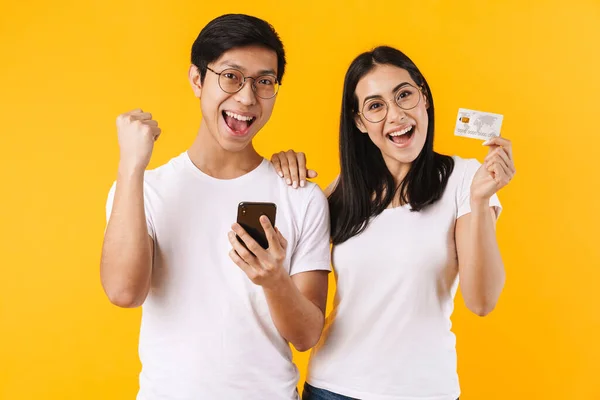 黄色の背景に隔絶されたカジュアルな服装をした美しい陽気な若いアジアのカップルの肖像画 携帯電話を保持し クレジットカードを示す — ストック写真
