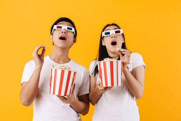 Image of shocked multinational couple in 3d glasses eating popcorn while watching movie isolated over yellow background