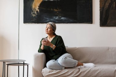 Beyaz saçlı, odaklanmış bir kadının oturma odasında koltukta otururken cep telefonu kullanmasının fotoğrafı.