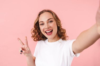 Pembe duvar arkasında izole edilmiş poz veren sevimli, iyimser genç bir kadının fotoğrafı. Fotoğraf çek ve barış jesti yap..