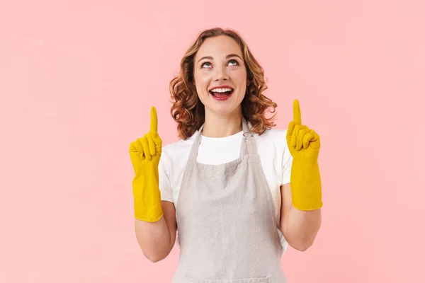 Photo of a cute positive young pretty woman housewife posing isolated over pink wall background in gloves pointing.
