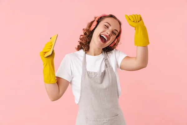 Stock image Photo of happy beautiful woman housewife posing isolated over pink wall background in gloves listening music with headphones holding phone.