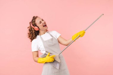 Duygusal ev hanımı görüntüsü pembe duvar arkasında eldivenlerle izole edilmiş. Elinde süpürgeyle gitar çalıyor gibi. Tonlarca müzik dinliyor..