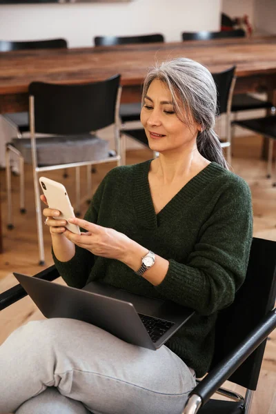 ラップトップコンピュータと携帯電話を使って屋内で成熟した素晴らしい幸せな女性のイメージ — ストック写真