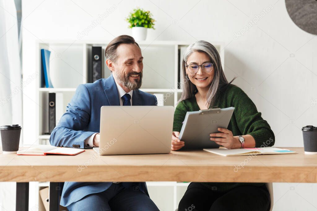 Photo of adult smiling colleagues talking while working with laptop and documents in office