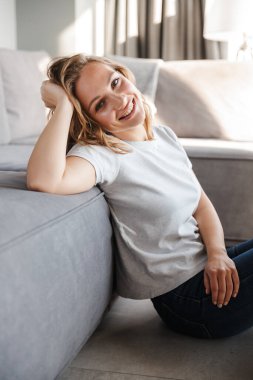 Oturma odasında yerde otururken kameraya gülümseyen beyaz kadın resmi.