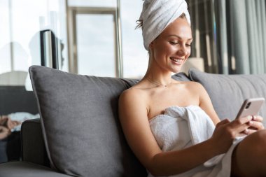 Duş aldıktan sonra odada otururken beyaz havlulara sarılı genç bir kadının fotoğrafı.