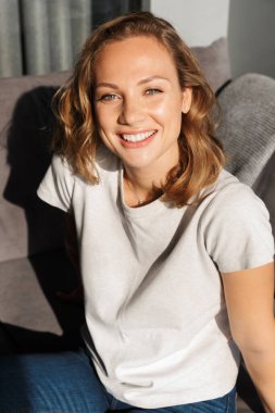 Gülümseyen, güzel, sarışın, günlük kıyafetler giyen, evdeki kanepede dinlenen genç bir kadın.