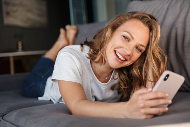 Oturma odasındaki kanepede yatarken gülümseyen ve cep telefonu kullanan basit tişörtlü mutlu sarışın kadın görüntüsü.