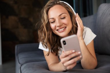 Oturma odasındaki kanepede uzanırken cep telefonu ve kablosuz kulaklık kullanan gülümseyen sarışın kadın.