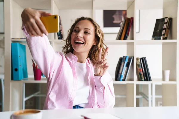 Foto Uma Jovem Sorrindo Tirando Selfie Celular Gesticulando Sinal Paz — Fotografia de Stock