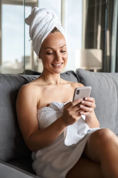 照片中 年轻快乐的女人在洗澡后坐在房间里 用白色毛巾包裹着手机 — 图库照片