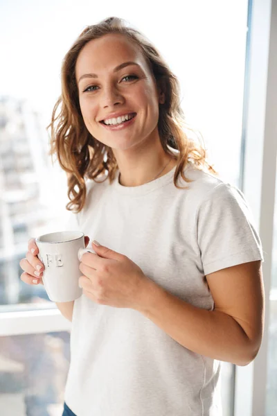 微笑的年轻女子站在室内的窗前 端着一杯茶 看着相机 — 图库照片