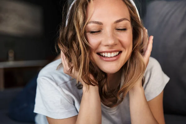 Obraz Uśmiechniętej Blondynki Używającej Bezprzewodowych Słuchawek Leżącej Kanapie Salonie — Zdjęcie stockowe