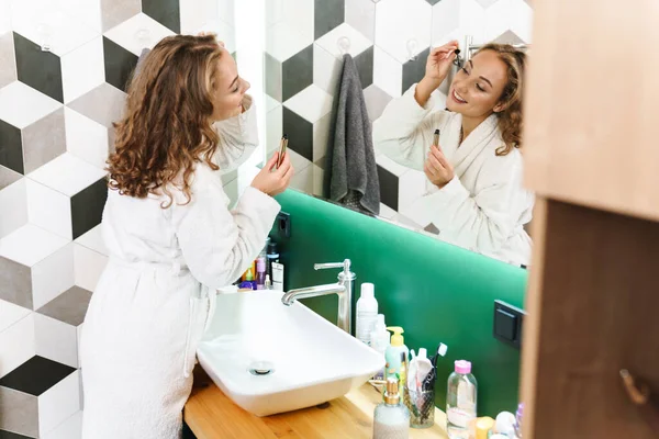穿着浴衣的年轻女子站在浴室的镜子前化妆 涂睫毛膏 — 图库照片