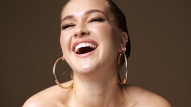 Harika, mutlu, yarı çıplak, şık küpeler takan bir kadın stüdyoda izole edilmiş bej arka planda kameraya gülüyor.