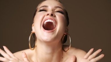 Şaşırmış, yarı çıplak, şık küpeler takan genç bir kadın, stüdyoda bej rengi arka planda izole bir şekilde mutlu oluyor.