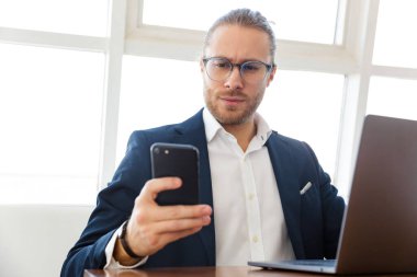 Ciddi genç iş adamının ofiste dizüstü bilgisayar ve cep telefonu kullanarak görüntüsü.