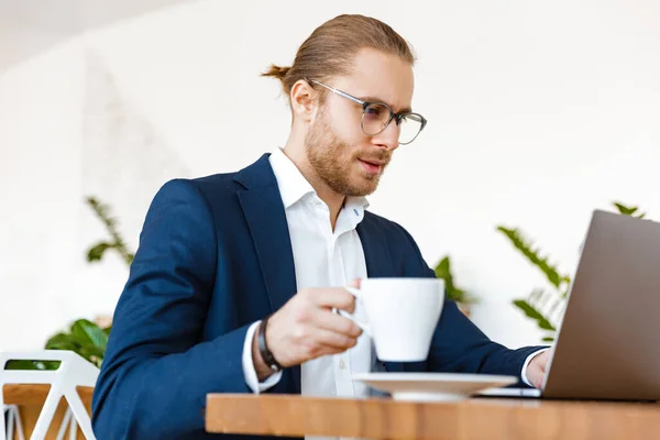 Yakışıklı Ciddi Genç Adamının Ofiste Dizüstü Bilgisayarla Kahve Içerken Görüntüsü — Stok fotoğraf