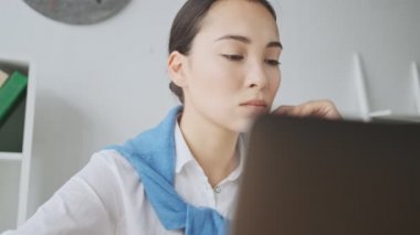 Asyalı düşünceli bir sekreter kadın ofiste çalışırken dizüstü bilgisayarını kullanıyor.